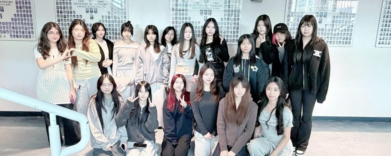 This image captures a group of approximately fifteen young women posing for a photo in what appears to be a school hallway. The group is arranged in two rows: a front row of seven women kneeling on the floor and a back row of eight women standing. They are all looking toward the camera, with various playful or neutral expressions. Several individuals are making hand gestures, such as peace signs or heart shapes, adding a casual and cheerful vibe to the photo.