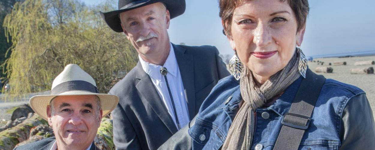 The image is a medium shot of three people posing outdoors on a beach on a sunny day. In the foreground, on the left, is a man wearing a light-colored fedora with a dark band, a grey blazer over a light blue collared shirt, and a brown guitar strap across his chest. He is looking directly at the camera with a slight smile and has short, dark hair.