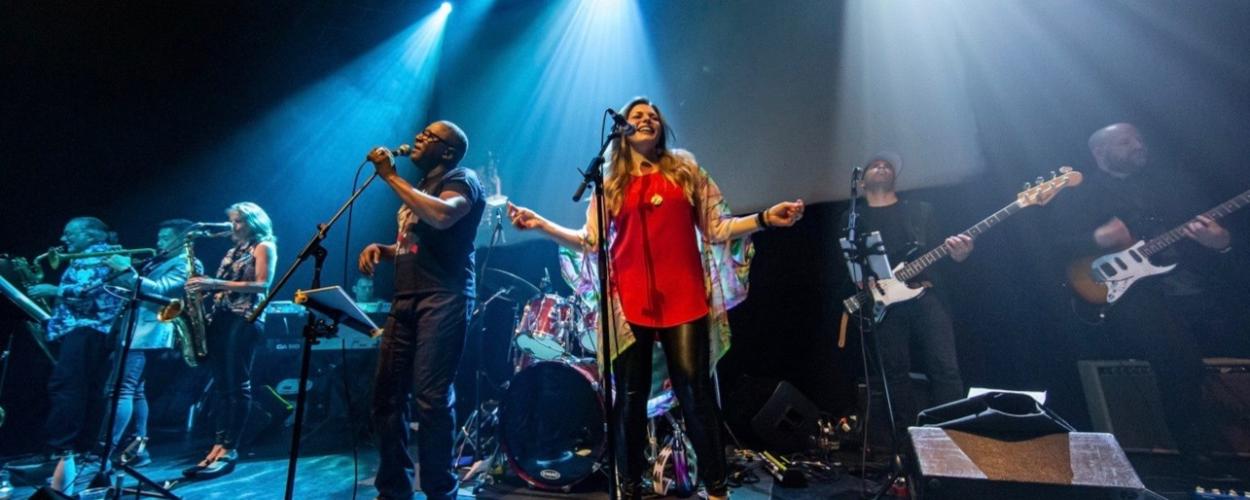 The image captures a live musical performance on a dimly lit stage. The primary focus is on a band with several musicians, illuminated by bright spotlights from above. The stage floor appears to be a dark, textured material, possibly wood or concrete, with some cables and equipment visible.