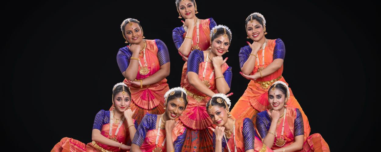 The image displays a group of eight young women performing what appears to be a traditional Indian dance. They are all dressed in vibrant, ornate costumes. Each dancer wears a blue, intricately patterned blouse with short sleeves and a full, orange skirt that has pink accents and pleats. Their attire is further adorned with heavy gold jewelry, including necklaces, earrings, bangles, and waistbands. They also wear decorative hairpieces, often with white elements. All the women have their hair styled up and a