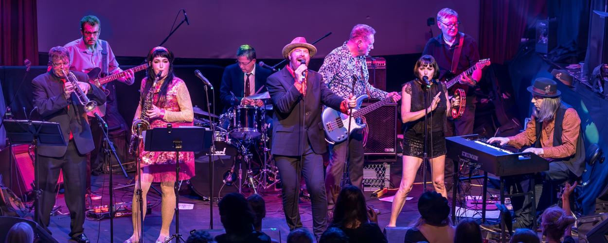 This is a medium shot of a live band performing on stage, captured under dramatic purple and blue stage lighting. The band consists of at least eight members, all actively engaged in their musical performance.