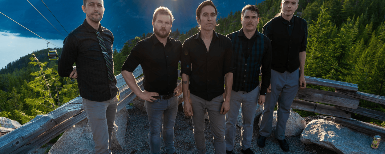 The image captures a group of five men standing on a rocky, gravelly overlook, seemingly in a mountainous region. The sun is setting or rising, casting a strong golden light with prominent sunbeams radiating from the top center of the frame, illuminating the scene