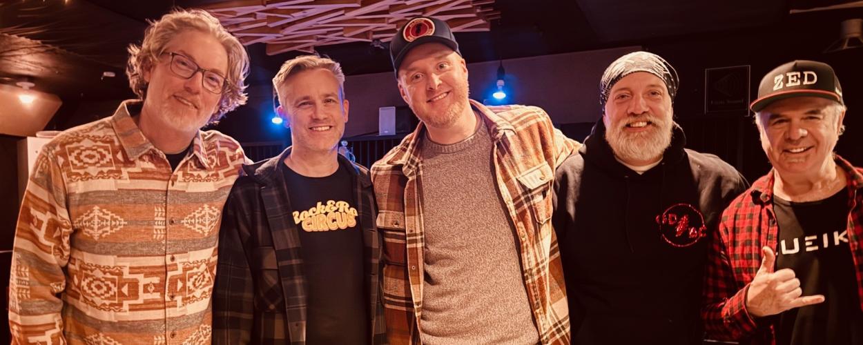 The image captures a group of five men posing for a photograph in what appears to be a dimly lit indoor space, possibly a studio or performance venue. The composition is a medium shot, focusing on the upper bodies of the individuals. They are arranged in a line, facing the camera with broad smiles, suggesting a friendly and relaxed atmosphere.