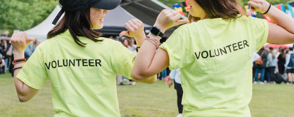 Two women in volunteer shirts smiling together in a park, surrounded by greenery and community spirit.