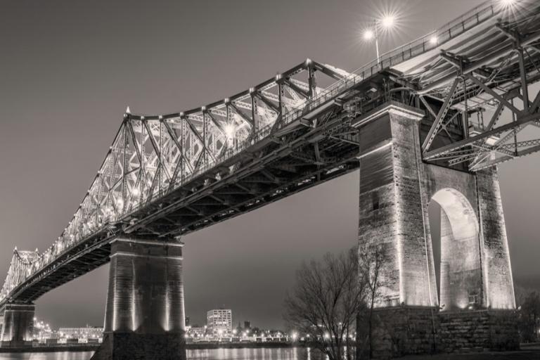 The image is a black and white photograph of a large, ornate bridge at night, illuminated by numerous lights. The perspective is from below and to the side, looking up at the imposing structure.