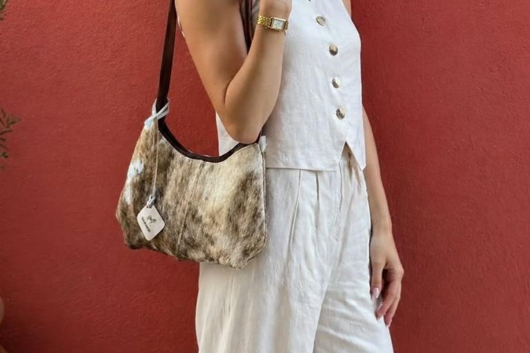 The image displays a medium shot of a person from the waist down, standing in front of a textured reddish-orange wall. The person is wearing a light-colored, possibly linen, outfit consisting of a sleeveless vest with four visible buttons and matching wide-leg pants. The vest is open, revealing a hint of the fabric underneath. The person is accessorized with a gold watch on their left wrist.