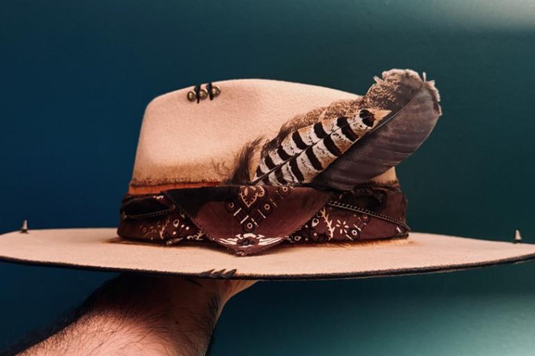 The image is a medium close-up, eye-level shot of a uniquely decorated hat being held by a hand. The hat is the central focus of the image. It appears to be a light tan or beige fedora-style hat with a moderately wide brim. The hat is adorned with several decorative elements. Around the base of the crown, there is a band made of dark reddish-brown fabric, possibly a bandana, with a paisley or similar intricate pattern in a lighter shade. The bandana is tied in the front, creating a visual knot and flowing e