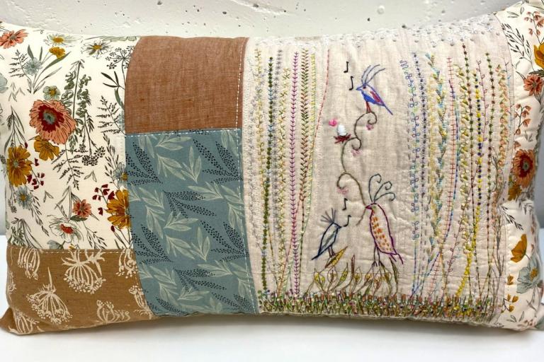 This is a close-up, eye-level shot of a rectangular, quilted decorative pillow. The pillow is oriented horizontally and is set against a plain white background, likely a wall. The pillow is divided into several panels with different fabric patterns and embroidered details.