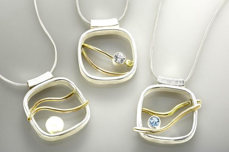 The image displays three contemporary silver pendants, each suspended from a fine, flexible silver chain. The pendants share a consistent design: a rounded square frame with a textured, brushed silver finish at the top where the chain attaches. Within each frame, a wavy, polished gold element is incorporated, adding a warm contrast to the cool silver. Each pendant also features a small, inset gemstone or pearl, positioned within the golden waves.