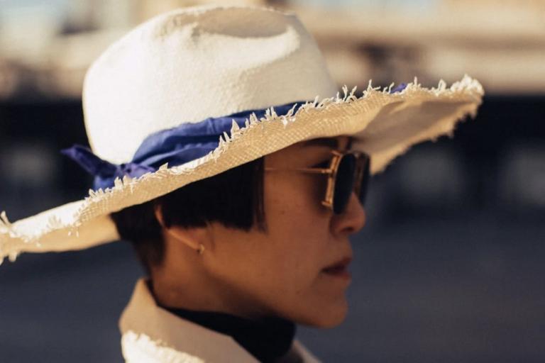 The image is a close-up, profile shot of a person wearing a wide-brimmed straw hat. The hat is white with a frayed edge and is adorned with a vibrant blue ribbon tied into a bow. The person, seen from the right side of their face, has short, dark hair with a blunt cut that peeks out from under the hat. They are wearing stylish gold-rimmed sunglasses with dark lenses, and a glimpse of a dark turtleneck is visible beneath the collar of a light-colored jacket or coat.
