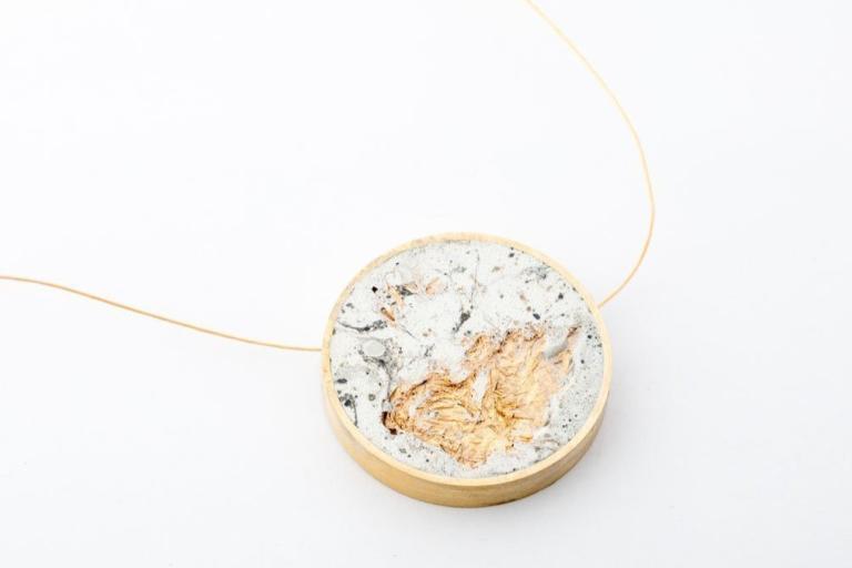 A close-up, high-angle shot features a circular pendant suspended from a delicate, thin gold chain. The pendant itself is the primary focus and is circular in shape, bordered by a polished gold rim that has a warm, slightly muted tone. The surface of the pendant is textured and appears to be made from concrete or a similar material, exhibiting a marbled pattern of light grey, white, and dark grey or black specks and swirls. Embedded within this concrete matrix is a prominent piece of textured, metallic gold