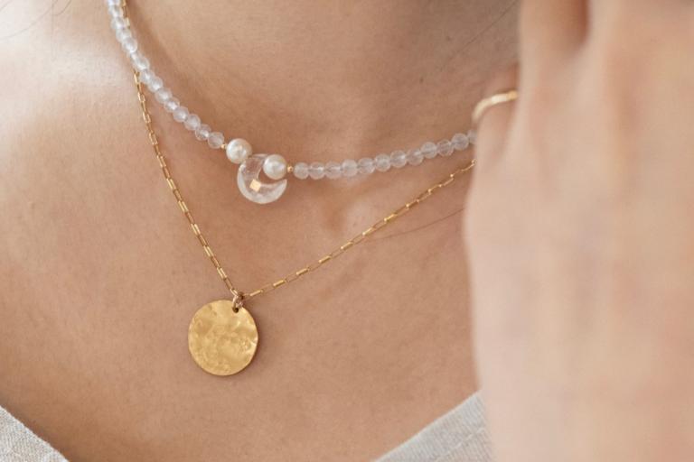 This is a close-up, waist-up, indoor shot featuring a person wearing two layered necklaces. The lighting is soft and natural, creating a gentle glow and highlighting the details of the jewelry. The background is out of focus, emphasizing the subject.