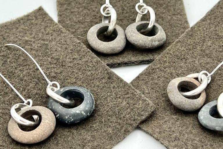 The image displays four pairs of handmade earrings, each featuring a smooth, weathered stone disk with a hole through its center, suspended from a delicate silver hook. The stones vary in color and texture, ranging from a light sandy beige to a darker, mottled grey and a reddish-brown hue. Some stones show subtle speckling and natural imperfections, adding to their unique charm. Each stone is attached to a silver hook with a single, slightly curved silver loop.