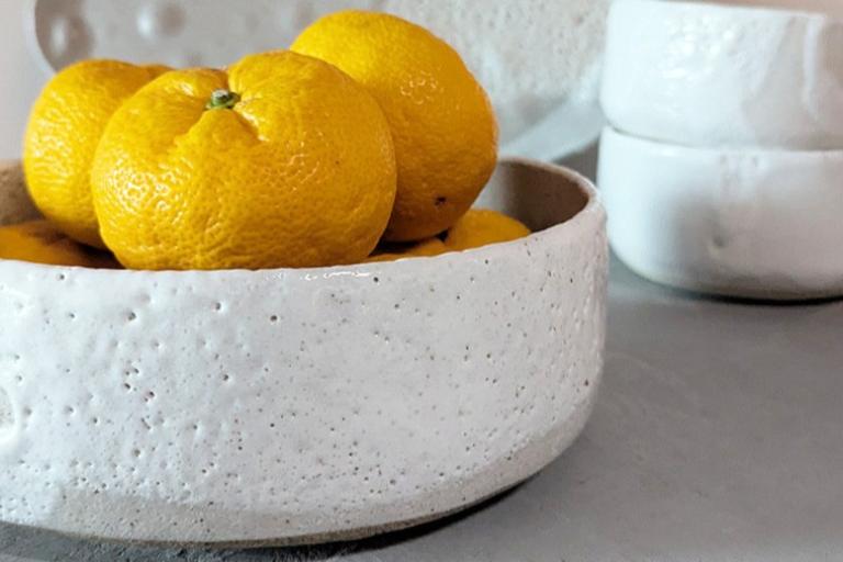 The image is a close-up, still-life shot focused on a rustic, textured white ceramic bowl filled with several bright yellow oranges. The bowl itself has an uneven, almost pitted surface, with subtle indentations and a rough-hewn rim. A circular impression is visible on the left side of the bowl's exterior. The oranges, clustered together, have slightly textured skins and appear plump and ripe. Their vibrant yellow hue stands out against the neutral tones of the pottery.