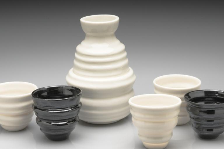 The image displays a collection of ceramic sake vessels arranged on a reflective surface against a gradient gray background. The central and largest object is a tall, ribbed sake bottle with a bulbous base that tapers into a narrow neck and a small opening. The bottle is a creamy white color and exhibits a series of horizontal ridges running down its body, creating a textured, layered appearance.