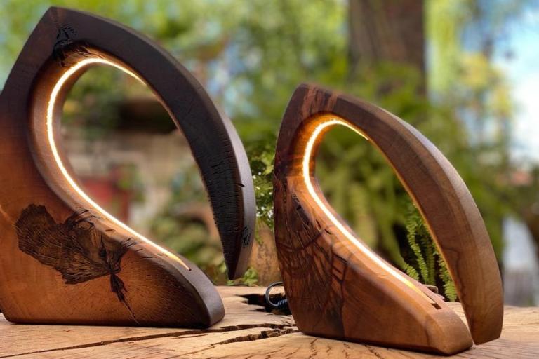 The image displays two striking, modern desk lamps crafted from wood, set against a softly blurred outdoor backdrop of lush greenery and hints of vibrant pink flowers. The lamps share a similar curvilinear, abstract design, resembling a crescent moon or a stylized horn.