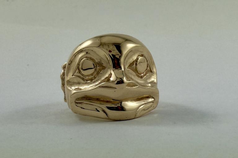 The image displays a close-up, front-facing view of a highly polished, gold-colored ring designed in the form of a stylized, possibly animalistic, head. The ring is centered within the frame, with the background being a plain, light, off-white surface that creates a soft, diffused illumination.