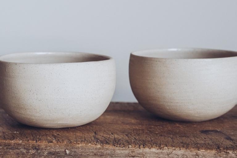 The image displays two handcrafted ceramic bowls placed side-by-side on a weathered wooden surface. The bowls are simple in design, with rounded bodies and slightly flared rims. Their color is a muted, earthy off-white with subtle variations in tone, suggesting they are unglazed or have a matte finish. Tiny dark specks are visible across the surface of both bowls, characteristic of stoneware clay.