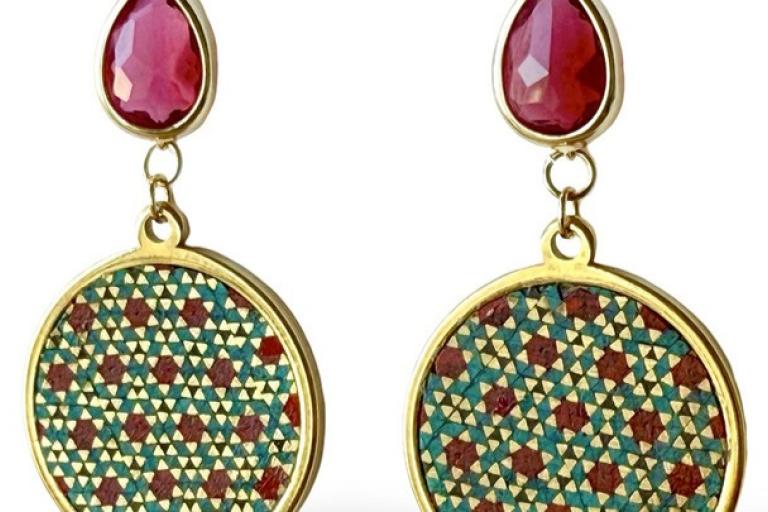 The image showcases a pair of exquisitely crafted dangle earrings against a stark white background. Each earring features a teardrop-shaped, faceted gemstone, likely a ruby due to its vibrant, deep red hue, set in a gleaming gold bezel. Suspended below this gemstone by delicate gold chains are circular pendants. These circular pendants are the focal point of the earrings' intricate design. 