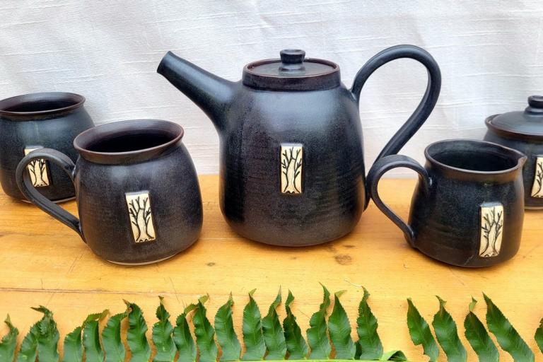 A rustic, handmade ceramic tea set is displayed on a wooden surface. The set includes a teapot, three mugs, and a lidded sugar bowl or creamer. All the pieces are a deep, matte black with a slight speckled texture, suggesting a natural clay body fired at a high temperature. Each item is adorned with a decorative rectangular plaque featuring a light-colored, stylized tree with bare branches. The interior rim of each mug and the teapot's lid and opening show a contrasting deep reddish-brown glaze.