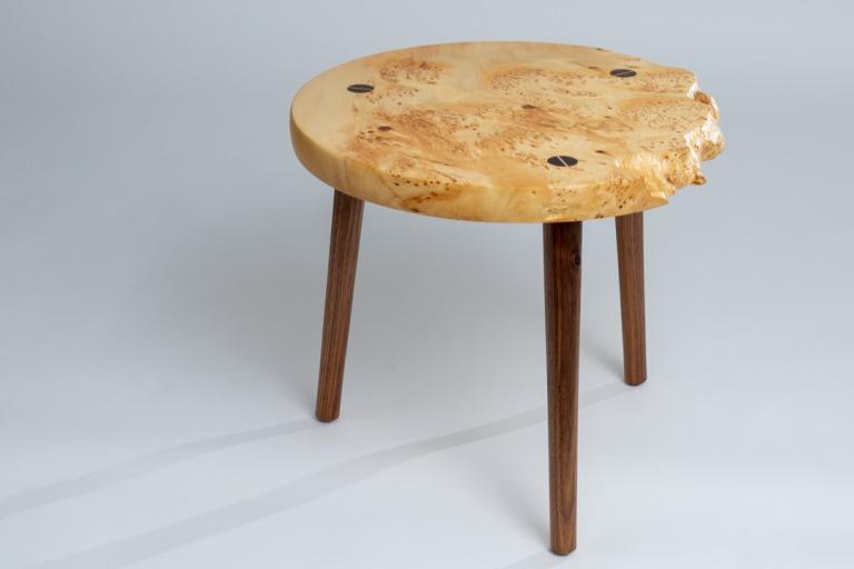 This is a studio shot of a unique, handcrafted wooden stool against a plain, light gray background. The stool is round and appears to be made from a single piece of light-colored wood, possibly burl wood, with a live edge on one side. The wood has a prominent grain pattern featuring swirls, knots, and darker speckles, giving it a very organic and textured look. Embedded into the surface of the stool are four dark, circular inlays, possibly made of a contrasting wood or a resin, arranged in a roughly rectang