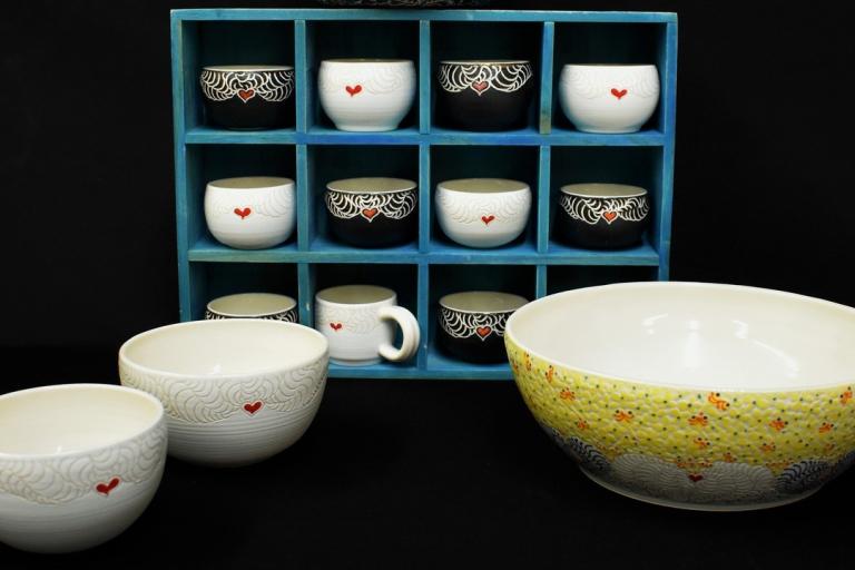 The image displays a collection of ceramic bowls and a mug, arranged against a plain black background. The primary focus is on the display of these handcrafted items, with a prominent blue wooden shelving unit holding several smaller pieces. The shelving unit is divided into twelve square compartments, arranged in three rows and four columns.