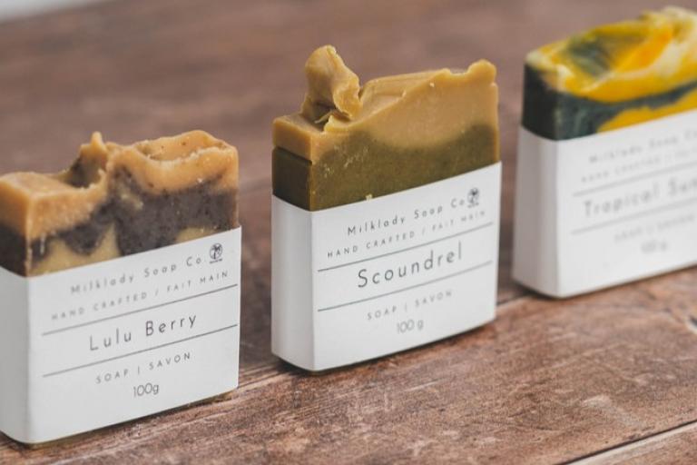 The image showcases three artisanal soap bars arranged in a diagonal line from left to right on a wooden surface. The soap bars are of varying heights and textures, with the leftmost bar being the shortest and the rightmost bar the tallest. Each soap bar is partially wrapped in a clean, white paper sleeve that displays product information.