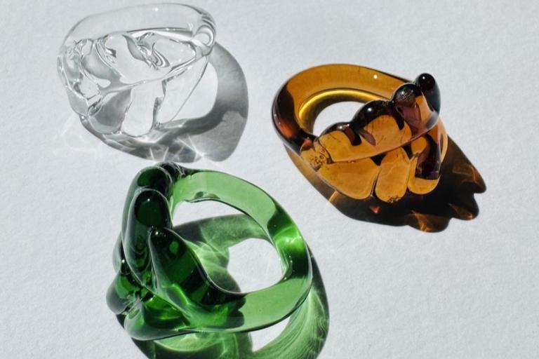 The image displays three chunky, transparent glass rings artfully arranged on a textured white surface. The rings are positioned in a triangular formation. The ring in the upper left corner is clear and colorless, with a fluid, organic shape that suggests it might be inspired by water or melting ice. Its surface reflects light with a slight opacity. To the right and slightly below this, a ring crafted from amber-colored glass is featured. This ring exhibits a more defined, almost floral or leaf-like pattern