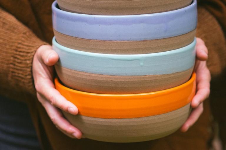 The image features a close-up shot of a stack of four ceramic bowls held by a person wearing a chunky, warm-toned brown knit sweater. The composition is centered on the bowls, with the hands and sweater acting as framing elements.