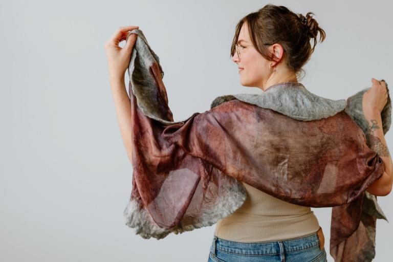 The image features a woman from the back, viewed from the right side. She is wearing glasses and her hair is pulled up in a bun. She is holding a large, flowing scarf around her shoulders. The scarf is a combination of colors, with a predominantly reddish-brown hue that appears to be dyed or naturally colored, possibly with botanical dyes. There are lighter, muted areas and darker, richer tones throughout the fabric, creating a mottled and organic pattern.