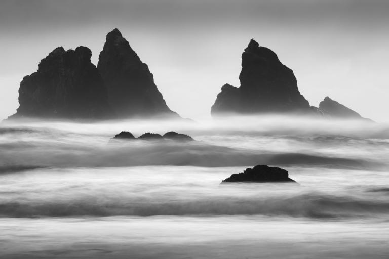 This is a black and white long exposure photograph of a dramatic coastal landscape. The scene is dominated by dark, jagged rock formations rising out of the ocean. The largest rock formations are positioned in the background, with the two most prominent ones appearing like imposing silhouettes against the sky. The rock on the left is larger and more complex, with multiple peaks. To its right, a slightly smaller rock formation stands, with a single, sharp peak. Further to the right, another, smaller rock for