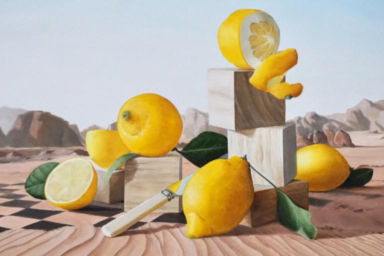 This is a surreal still life painting with a desert landscape in the background. The foreground is dominated by several lemons, arranged in a somewhat precarious composition. There are whole lemons, halved lemons, and a lemon peel. The lemons are a vibrant, saturated yellow, with some showing subtle variations in tone and texture. One lemon at the top is cut in half, revealing its juicy interior and segmented pulp. A spiral of lemon peel elegantly drapes over a stack of wooden blocks, which are themselves a