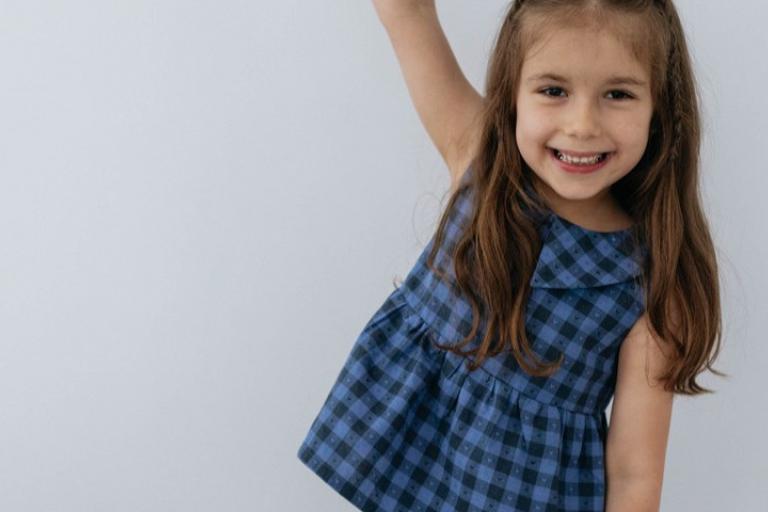 A young girl with long, brown hair is captured in a medium shot, smiling warmly at the camera. Her hair is parted in the middle, with two thin braids on each side framing her face, and the rest falling loosely down her shoulders. She is wearing a sleeveless blue dress with a subtle checkered pattern and a Peter Pan collar.