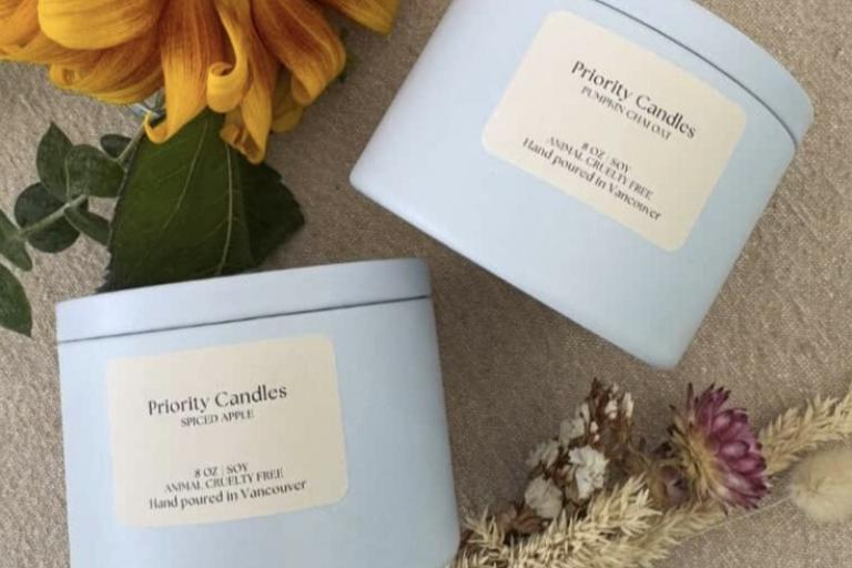 The image displays a close-up, overhead shot of two light blue tin candles placed on a textured, neutral-toned fabric surface. The candles are positioned diagonally, with one slightly above and to the right of the other. Both candles have cream-colored labels with text. The top candle's label reads "Priority Candles" in a prominent font, followed by "PUMPKIN CHAI OAT" in a smaller font. Below this, it states "8 OZ. SOY ANIMAL CRUELTY FREE Hand poured in Vancouver." The bottom candle has a similar label, wit