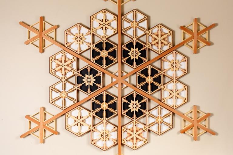 The image showcases a decorative wall hanging or artwork designed to resemble a snowflake. It is composed of numerous interlocking hexagonal shapes arranged in a symmetrical, radial pattern. The primary materials appear to be wood, with variations in color suggesting different types or finishes.