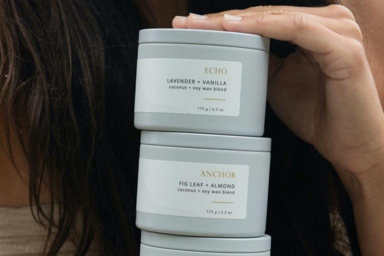 The image shows a close-up of a person's hands holding a stack of three light gray metal candle tins. The tins have minimalist white labels with thin gold line graphics and text. The top tin is labeled "ECHO" and "LAVENDER + VANILLA", and indicates a "coconut + soy wax blend" and a weight of "175 g | 6.2 oz.". The second tin from the top is labeled "ANCHOR" and "FIG LEAF + ALMOND", also with the coconut + soy wax blend and weight. A third tin is partially visible at the bottom of the stack. The person's han