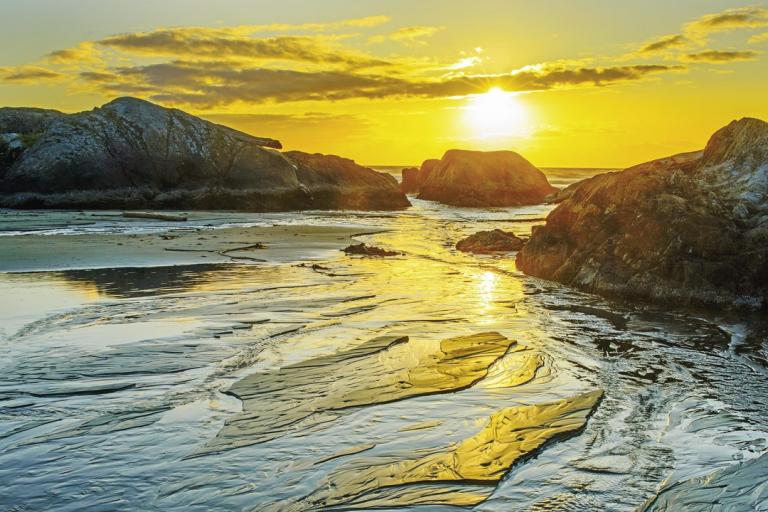 The image captures a serene coastal scene at sunset, characterized by vibrant golden light and natural textures. The sun, a brilliant orb of intense yellow and white, is positioned in the upper right quadrant of the frame, casting long, warm rays across the landscape. It's partially obscured by scattered clouds that are illuminated with bright yellow and orange hues, creating a dramatic sky.