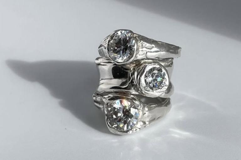 The image displays a stack of three silver rings, each adorned with a clear, faceted gemstone. The rings are arranged in a slightly offset, vertical stack, with the top ring positioned at an angle. The gemstones appear to be diamonds or a similar clear stone, reflecting light and showing internal facets. The silver bands of the rings have a rough, organic texture, suggesting a handcrafted or artisanal design, rather than a polished, uniform finish. The texture is uneven, with visible bumps and undulations.