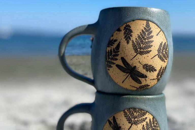 The image is a close-up shot of two stacked ceramic mugs, positioned in the foreground with a blurred background of a beach and ocean scene. The mugs are a muted blue-gray color with a speckled, matte finish. Each mug features a circular decal or inlay on its side. This circular design is a light tan color with a textured surface. Within these circles, there are black imprints of botanical elements, specifically fern fronds, and a prominent dragonfly. The dragonfly is depicted in flight, with its wings spre