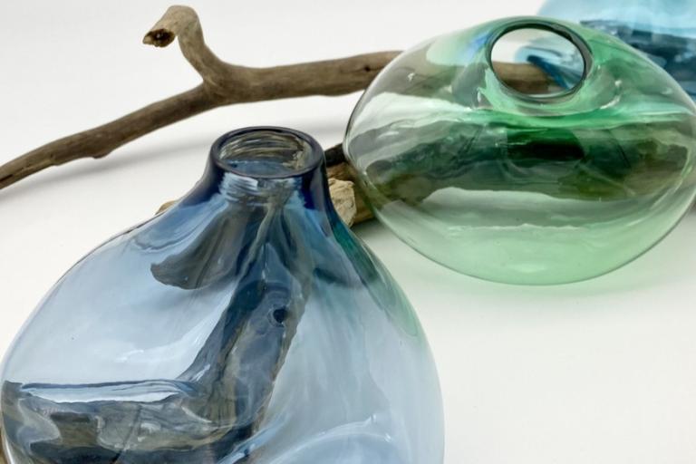 The image displays a still life composition featuring three blown glass vessels and pieces of driftwood, set against a plain white background. In the foreground, on the left, is a large, pear-shaped glass vessel with a neck. The glass is a gradient of deep blue at the neck transitioning to a lighter, almost translucent blue towards the base. Inside this vessel, a piece of driftwood is visible, appearing almost sculptural within the glass. The texture and color of the wood are clearly discernible through the
