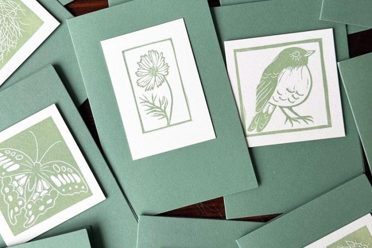 The image is a close-up, overhead shot of a collection of handmade cards with nature-themed linocut prints. The cards are arranged in a slightly overlapping, scattered manner on a dark, wooden surface, visible in the background between the cards.
