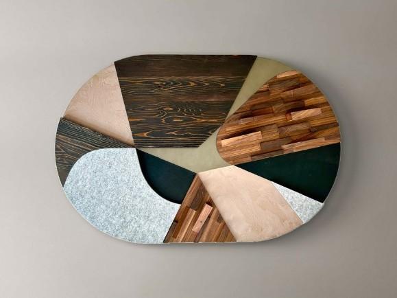 This image showcases an abstract wall art piece mounted on a neutral gray wall. The artwork itself is a large, oval-shaped composition with a textured, multifaceted surface. It's constructed from various materials and geometric shapes.