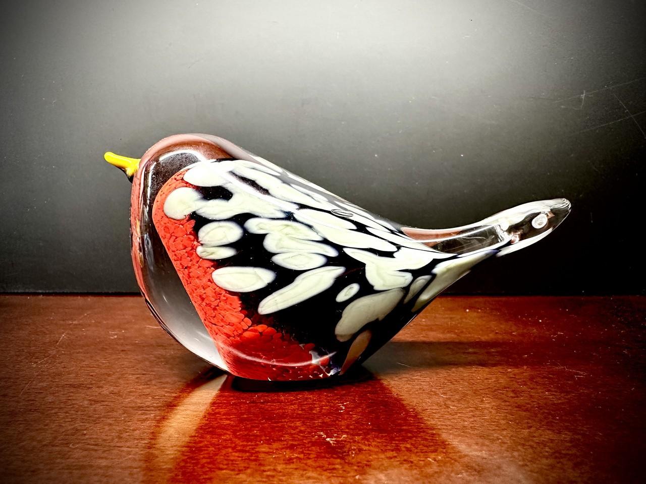 A glass figurine of a bird, resembling a robin, is positioned on a polished wooden surface. The figurine is made of clear glass with embedded colors and textures. The front of the bird, including its chest and part of its head, features a bright, textured red, reminiscent of granulated sugar or tiny beads. This red transitions into a dark, almost black, glass that forms the body and wings. Embedded within the black glass are irregular, elongated shapes of white and off-white, creating a speckled or feathere
