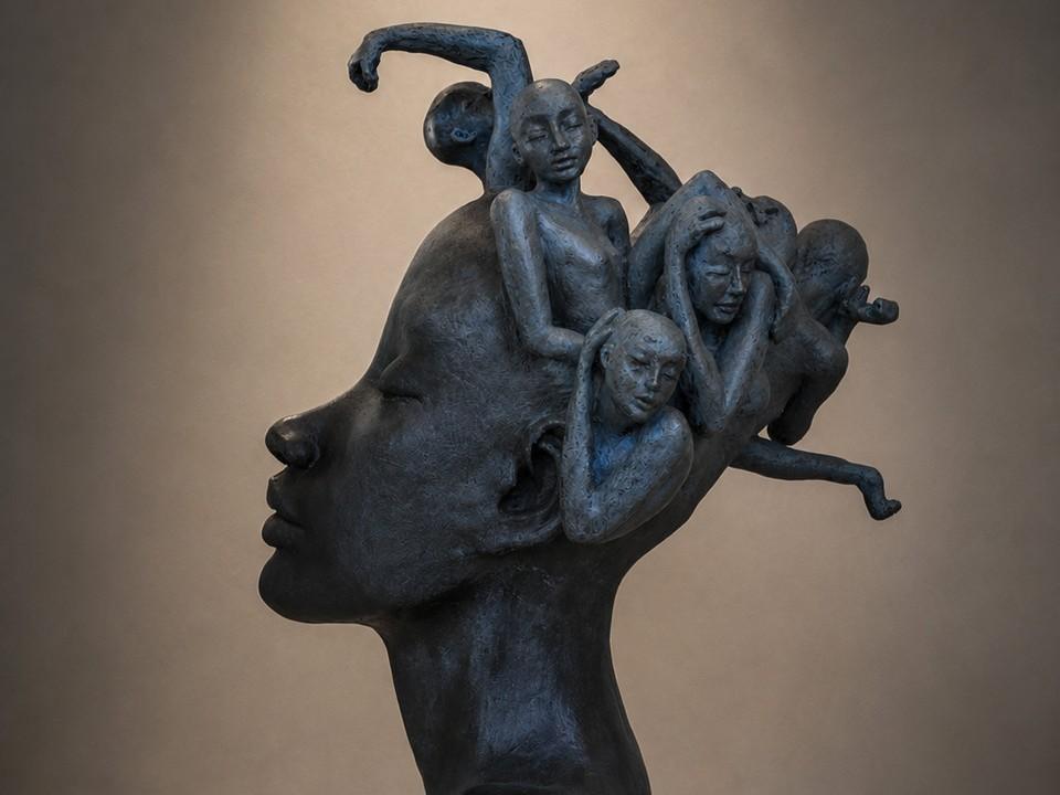 The image is a close-up, side-profile view of a dark gray, abstract sculpture. The sculpture depicts the head of a stylized human, seen from the left side with closed eyes and a neutral expression. Emerging from the top and back of this head are several smaller, intertwined human figures in various contorted poses. These smaller figures appear distressed or overwhelmed, with some clutching their heads or stretching their arms in what could be interpreted as anguish or struggle. 