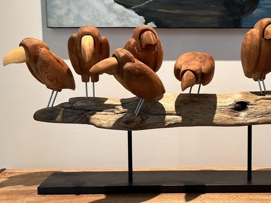 This image showcases a charming wooden sculpture of several stylized birds perched on a piece of driftwood. The birds are carved from a warm, reddish-brown wood, exhibiting visible grain patterns and a smooth, polished finish. Their forms are simplified and abstract, with round bodies and distinct, beaked heads. Some birds have lighter-colored beaks, adding a subtle contrast. They are supported by thin, silver metal rods that emerge from their undersides, attaching them to the driftwood.