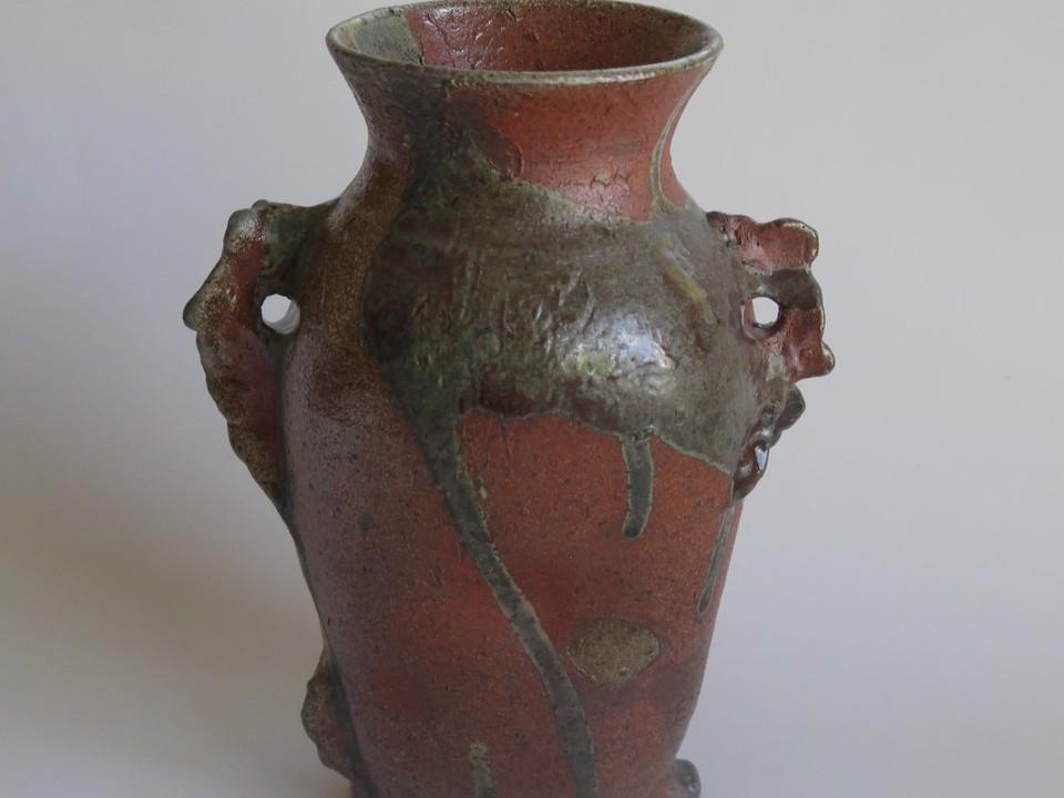 This is a medium close-up, eye-level shot of a ceramic vase. The vase has a somewhat irregular, organic shape with a narrow neck that flares out slightly at the rim. The dominant color of the vase is a rustic, deep reddish-brown, reminiscent of terra cotta. Streaked and dripped across the body of the vase is a glaze in muted, earthy tones of greyish-green and a lighter, almost yellowish-green. These drips create a dynamic, almost fluid appearance on the textured surface.