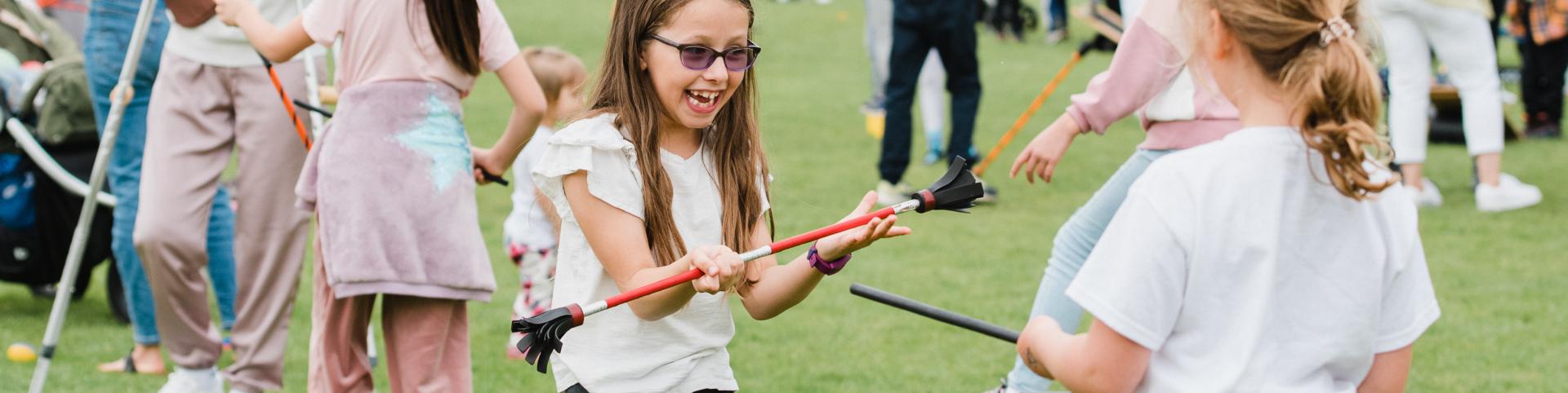 The image captures a lively and joyful outdoor scene where children are actively engaged in playful activities on a grassy field. In the foreground, a young girl with glasses grins excitedly as she plays with a foam-tipped stick alongside another child, both clearly enjoying the moment. Around them, other children and families participate in similar activities, creating a bustling, energetic atmosphere. The background is filled with a diverse crowd, adding to the vibrant community feel of what appears to be