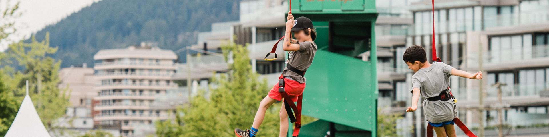 The image shows two children enjoying a bungee trampoline activity at an outdoor event. They're suspended mid-air, each harnessed and attached to red bungee cords connected to a tall green tower labeled "BNB Inflatables – Let the Fun Begin!" Both kids are wearing casual clothes and appear to be having a great time, with buildings and forested mountains in the background. There are white and green event tents in the foreground, suggesting this is part of a larger festival or community gathering in an urban p