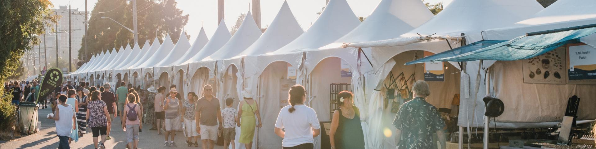 A bustling art market scene with a large group of people strolling along a street filled with colorful tents.