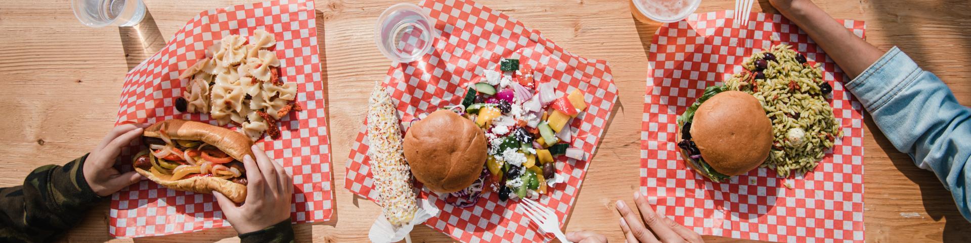 A picnic table is set with a variety of food, including a hot dog, a burger, a salad, and pasta salad. The food is placed on red and white checkered paper. There are also two glasses of water and a glass of beer. People are seen in the image, but only their hands and arms are visible.  They are enjoying their meal outdoors on a sunny day.